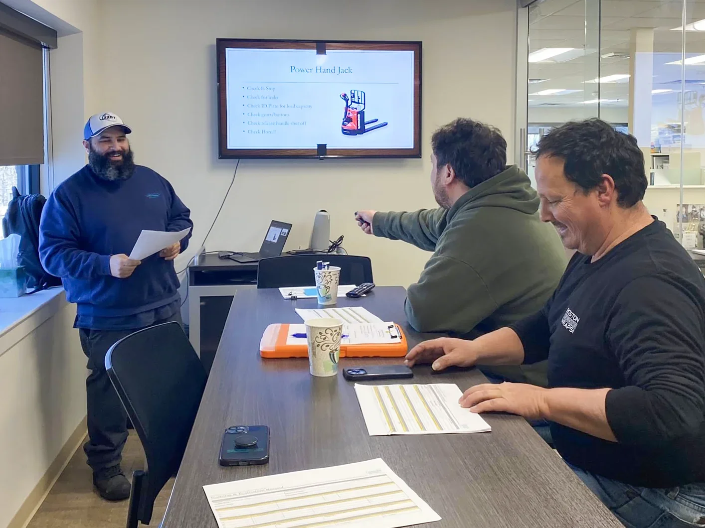 Three men in a meeting room with documents and coffee cups, one standing and presenting a PowerPoint slide titled 'Power Hand Jack,' which has a list of instructions and an illustration of a hand jack. Two men are seated at the table, smiling and engaging with the presentation.