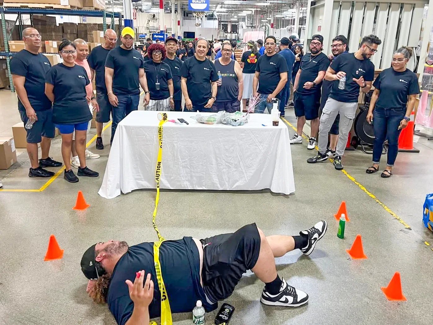 Group of coworkers in a warehouse setting standing behind a table, with a man lying on the floor in front of the table. The man on the floor appears to be participating in a safety demonstration or drill, with traffic cones and caution tape around him. Others are watching, some smiling or using their phones.