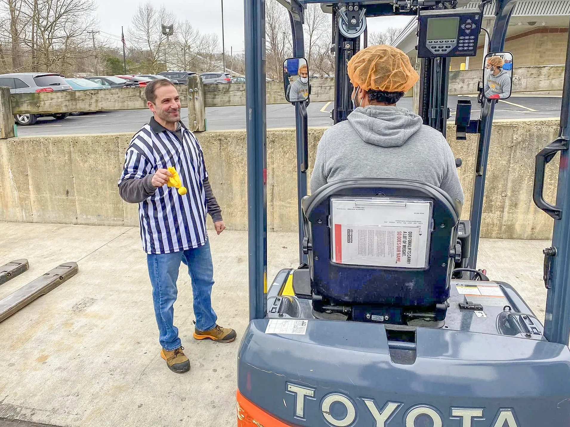 A man in a striped referee shirt holding a yellow balloon is standing outside, talking to a person operating a forklift. The forklift is marked with 'TOYOTA' and is parked next to a concrete wall. The person operating the forklift is wearing a gray hoodie and an orange hairnet.