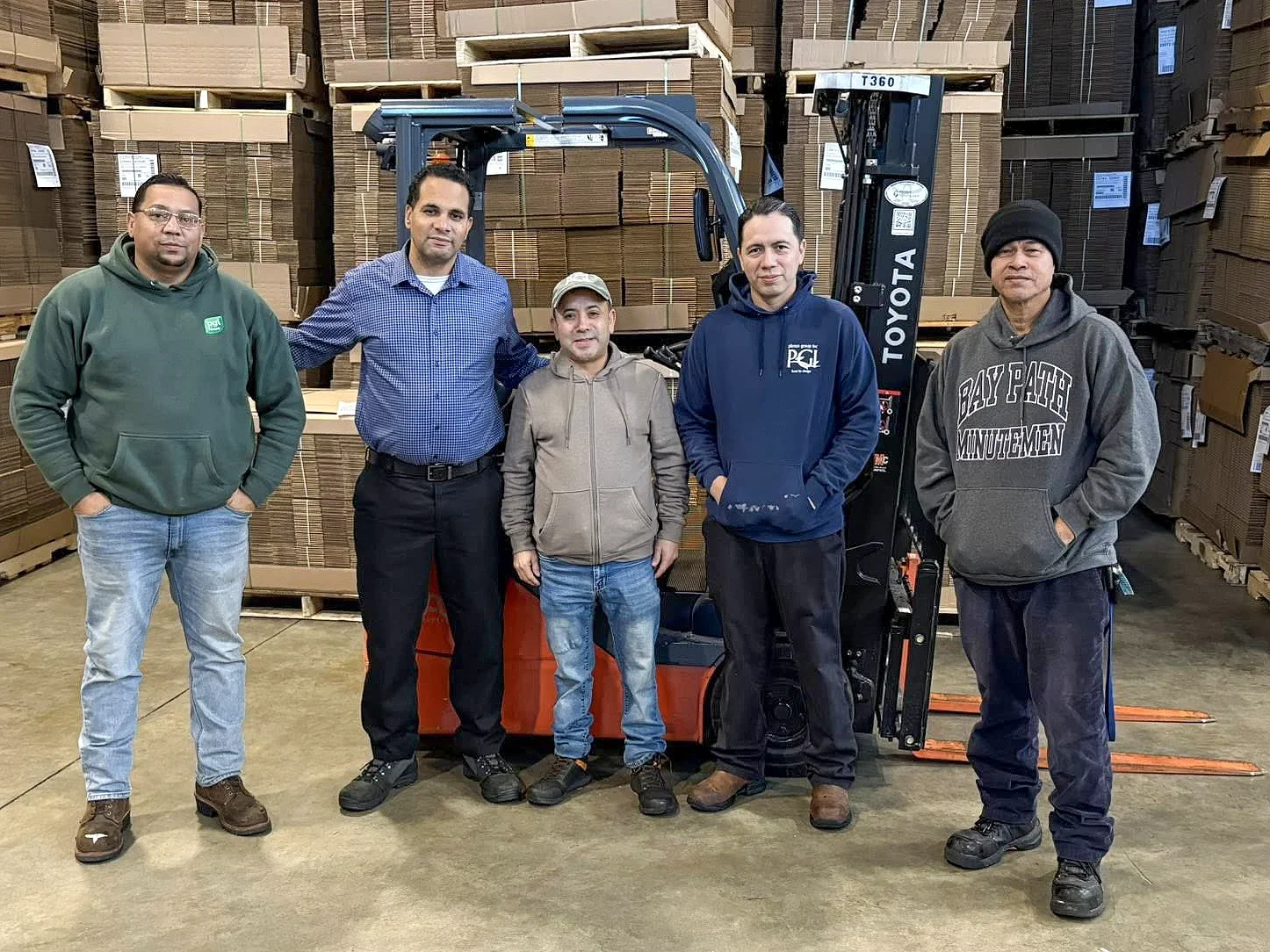 Five men standing in a warehouse with stacks of cardboard on shelves behind them, beside a forklift.