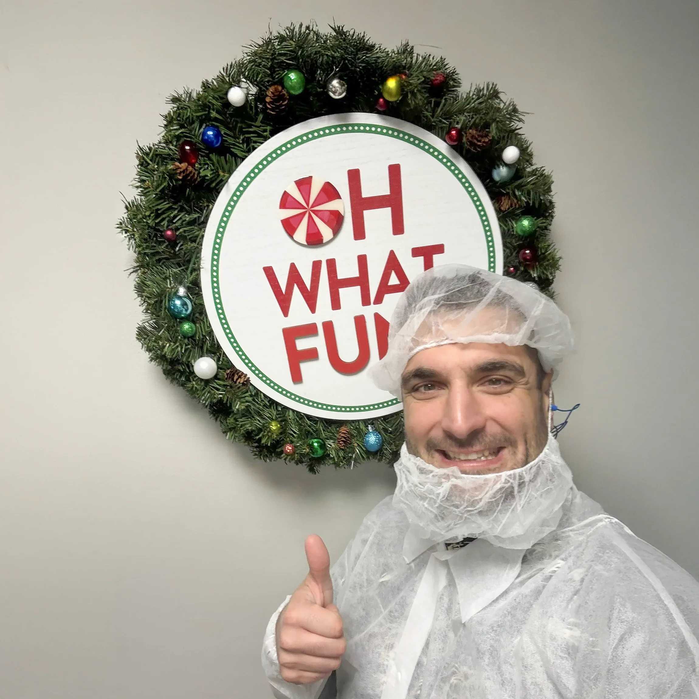 A smiling man wearing a hairnet and protective suit giving a thumbs up in front of a holiday wreath with a sign that says 'OH WHAT FUN' decorated with ornaments and pine cones.