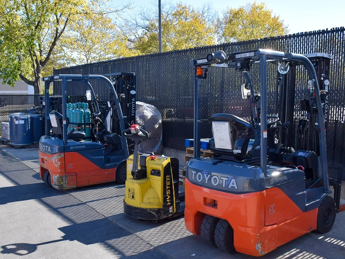 Two Toyota forklifts and a yellow pallet jack in an outdoor storage area with trees and black metal fencing in the background.