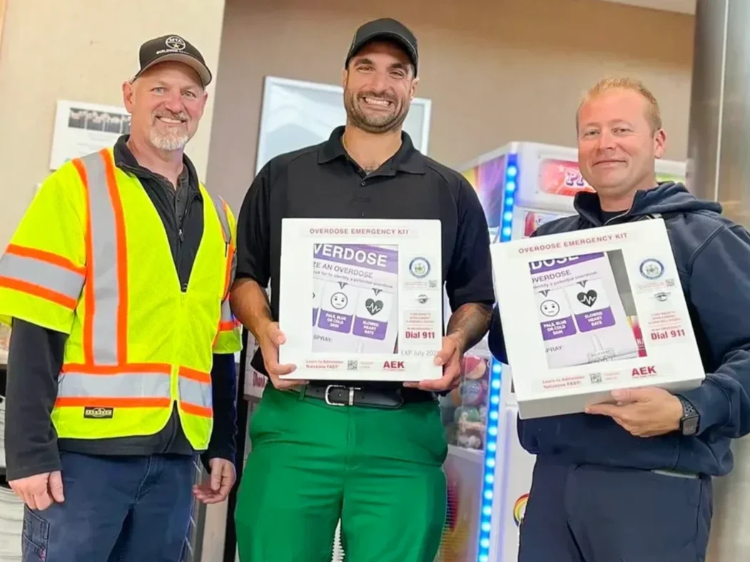 Three smiling men standing indoors, two of whom are holding boxes labeled 'Overdose Emergency Kit' containing overdose rescue supplies. The man on the left is wearing a high-visibility safety vest and a baseball cap. The middle man is dressed in a black polo shirt and green pants, and the man on the right is wearing a dark hoodie.