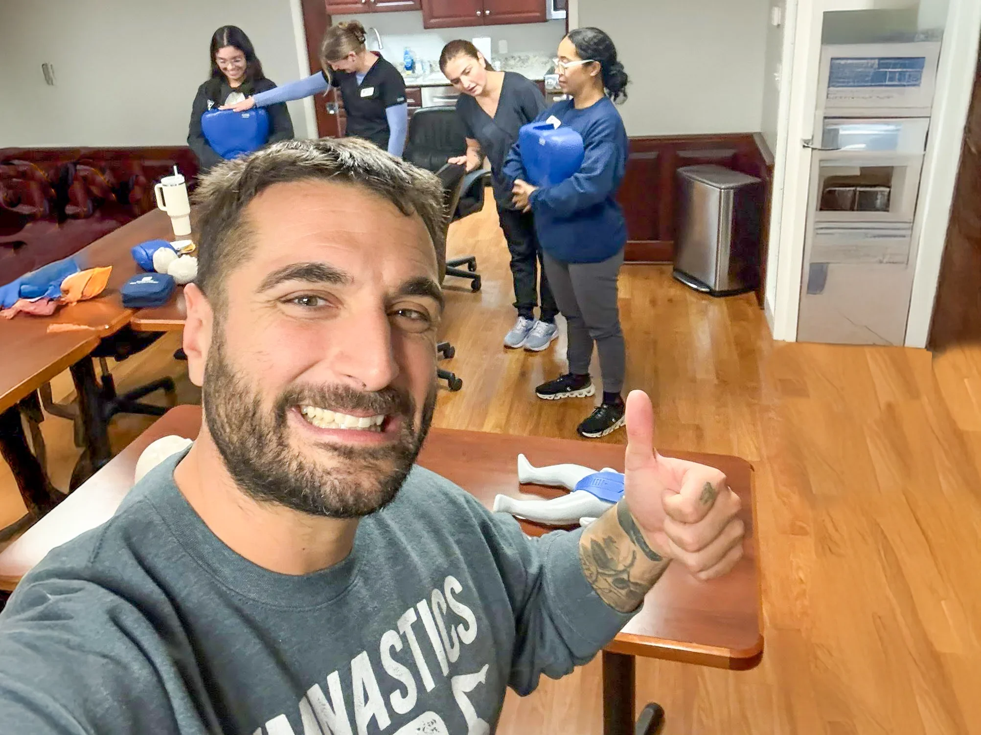 Man taking a selfie with four women in the background holding blue containers in a room with wooden floors and furniture.