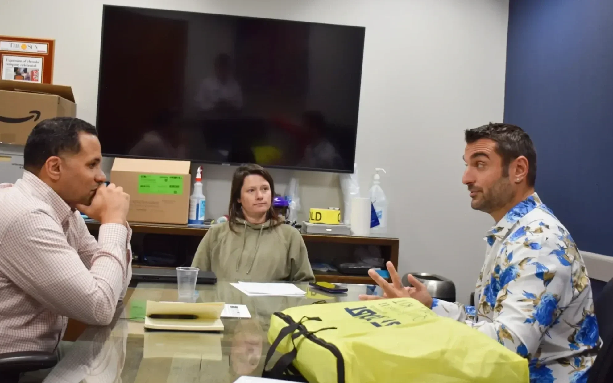 Three people sitting around a conference table in a room with a large television mounted on the wall. The person on the right is speaking, wearing a white shirt with blue floral pattern. The person on the left appears to be listening with their hand on their chin. The woman in the middle is looking at the speaker. There are various items on the table, including a yellow bag, a notepad, and some papers.