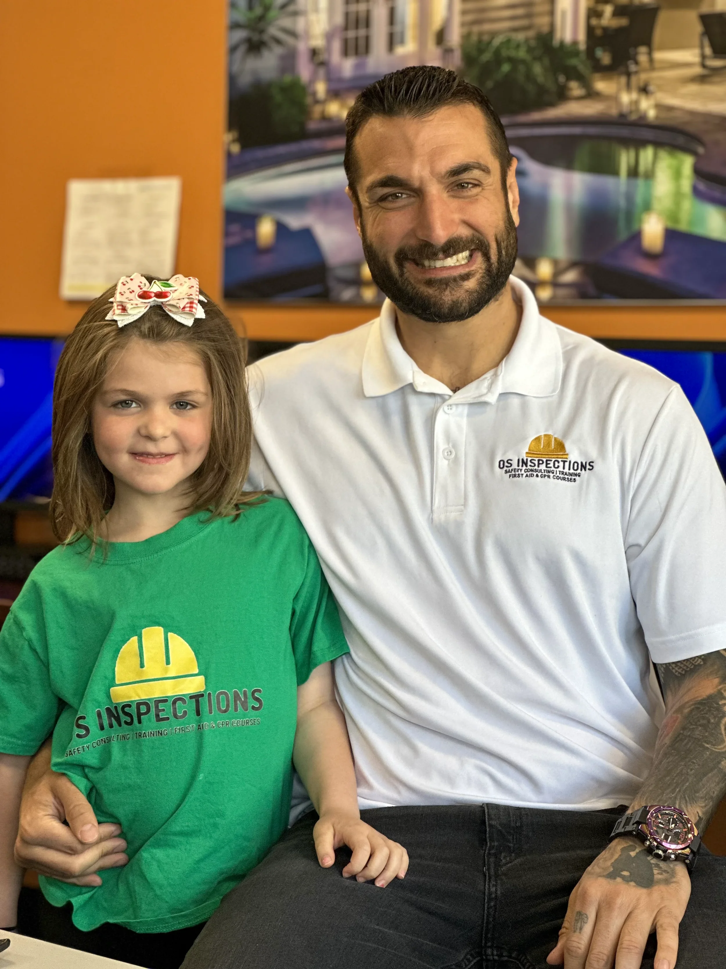 A man and a young girl sitting together, both wearing green and white shirts with a safety inspection logo. The girl has a bow in her hair, and they are smiling at the camera, indoors with a large screen displaying an outdoor scene in the background.