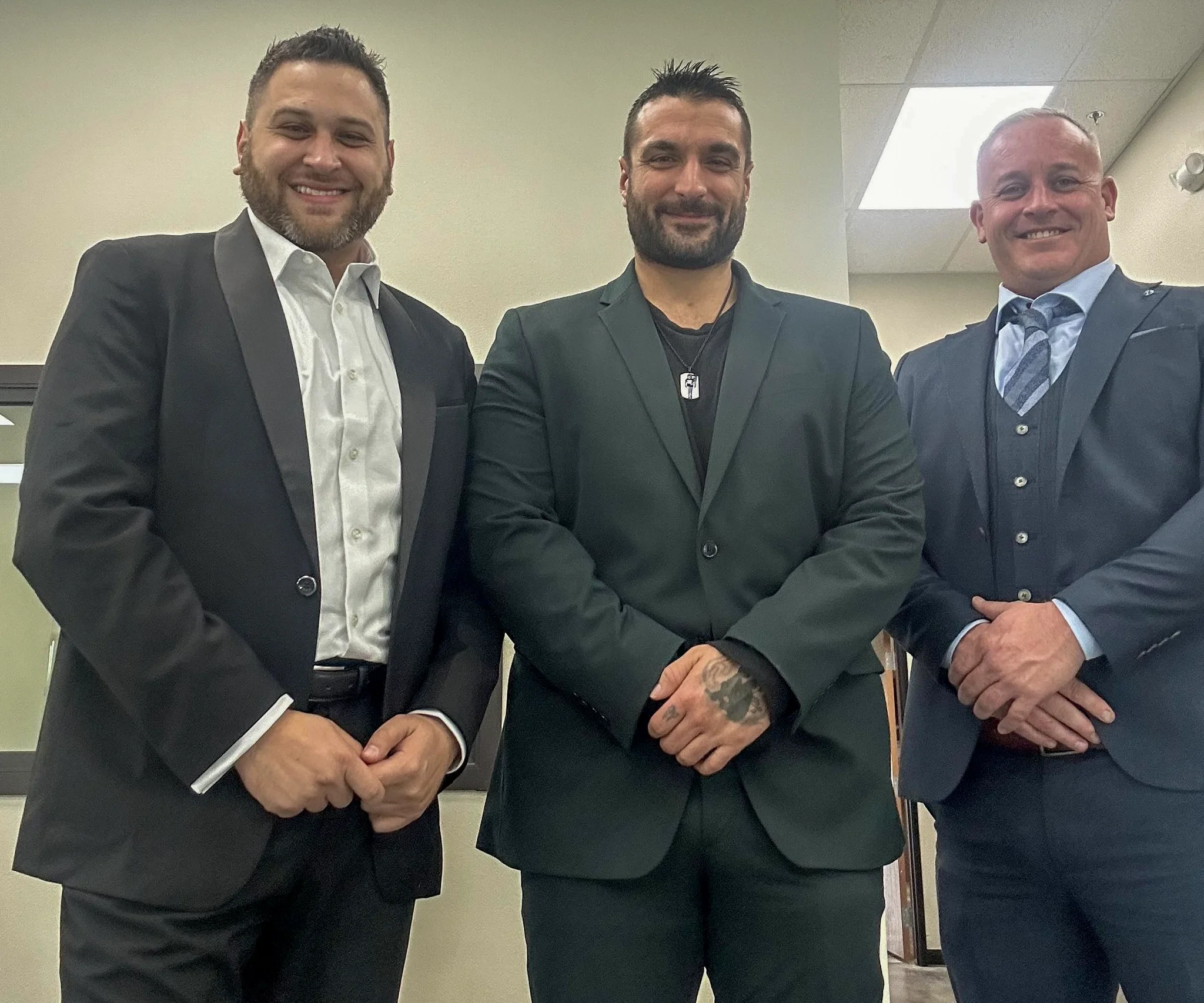 Three men in suits standing side by side indoors, smiling at the camera.