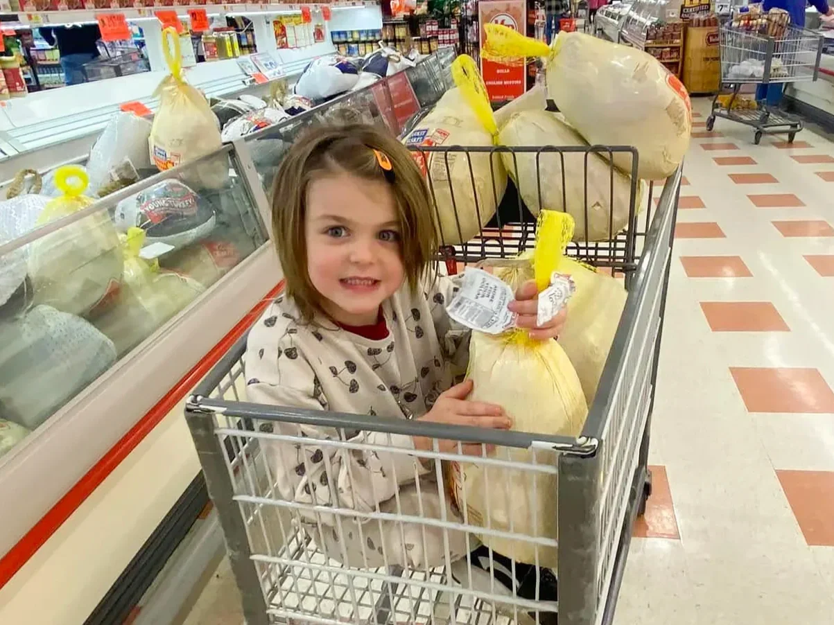 Young girl with shoulder-length brown hair inside a grocery cart, holding a large bag of food and a yellow item, in a grocery store aisle with frozen foods on the side.