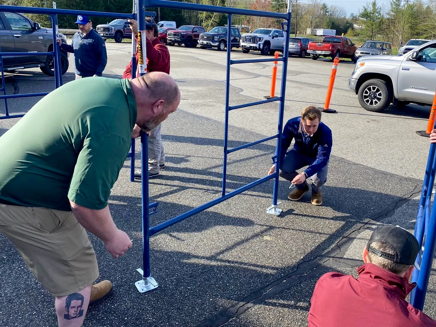 A group of people are assembling a blue metal frame outdoors in a parking lot. One man in a green shirt and khaki shorts is bending down, another man in a blue jacket is squatting, and others are standing nearby.