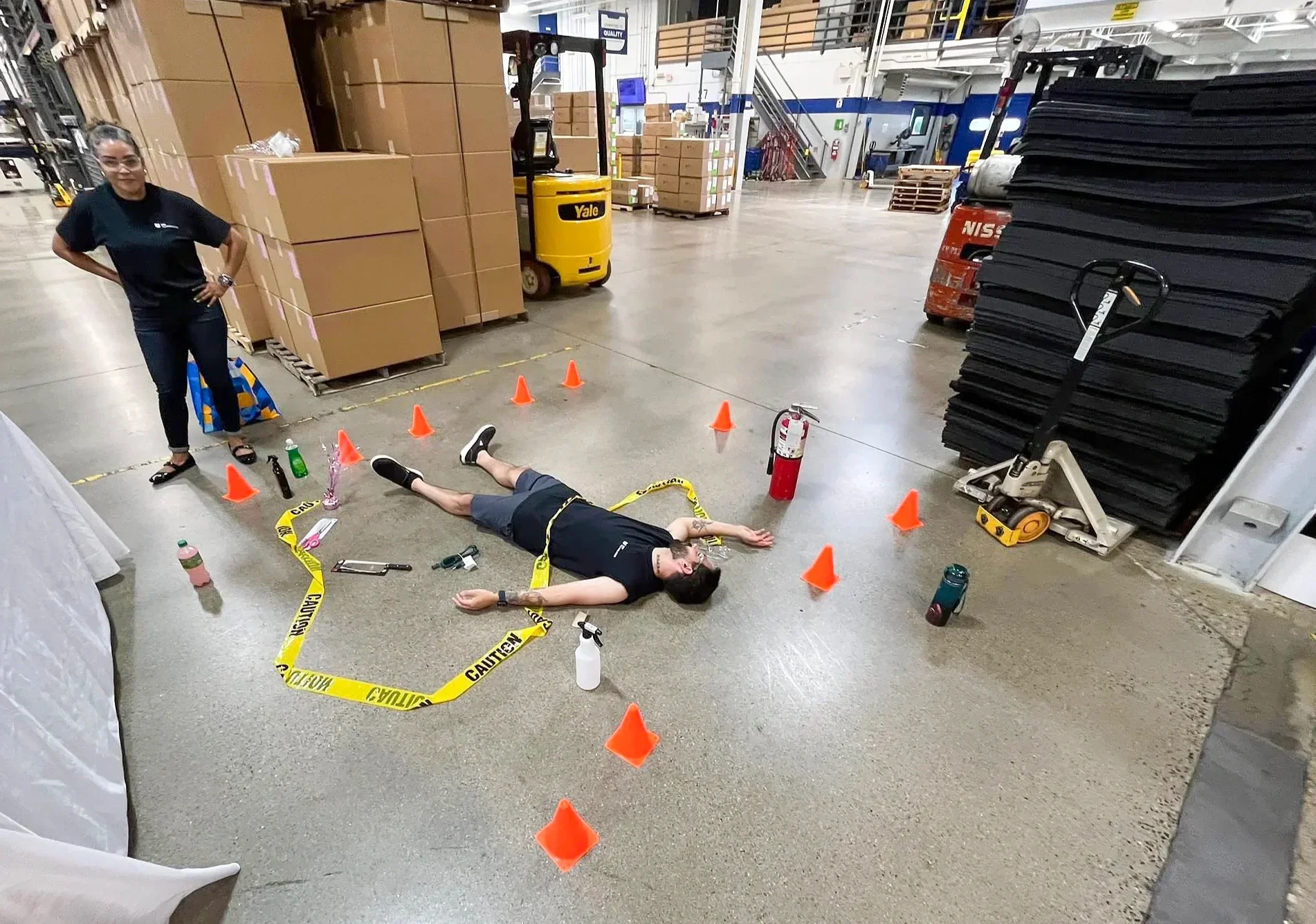 An indoor warehouse setting with a woman standing and a man lying on the floor surrounded by orange cones, caution tape, water bottles, and tools. The man appears to be participating in a safety drill or training exercise.