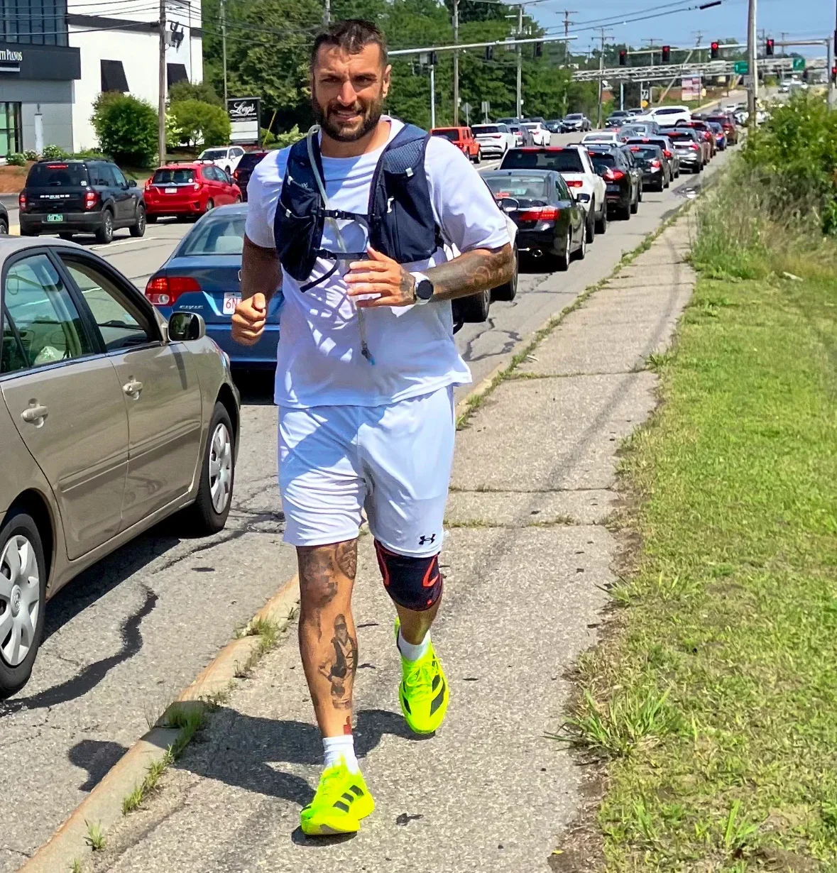 A man running on a sidewalk beside a busy street, dressed in athletic clothing, wearing neon yellow running shoes, with a backpack and a tattoo on his left leg.