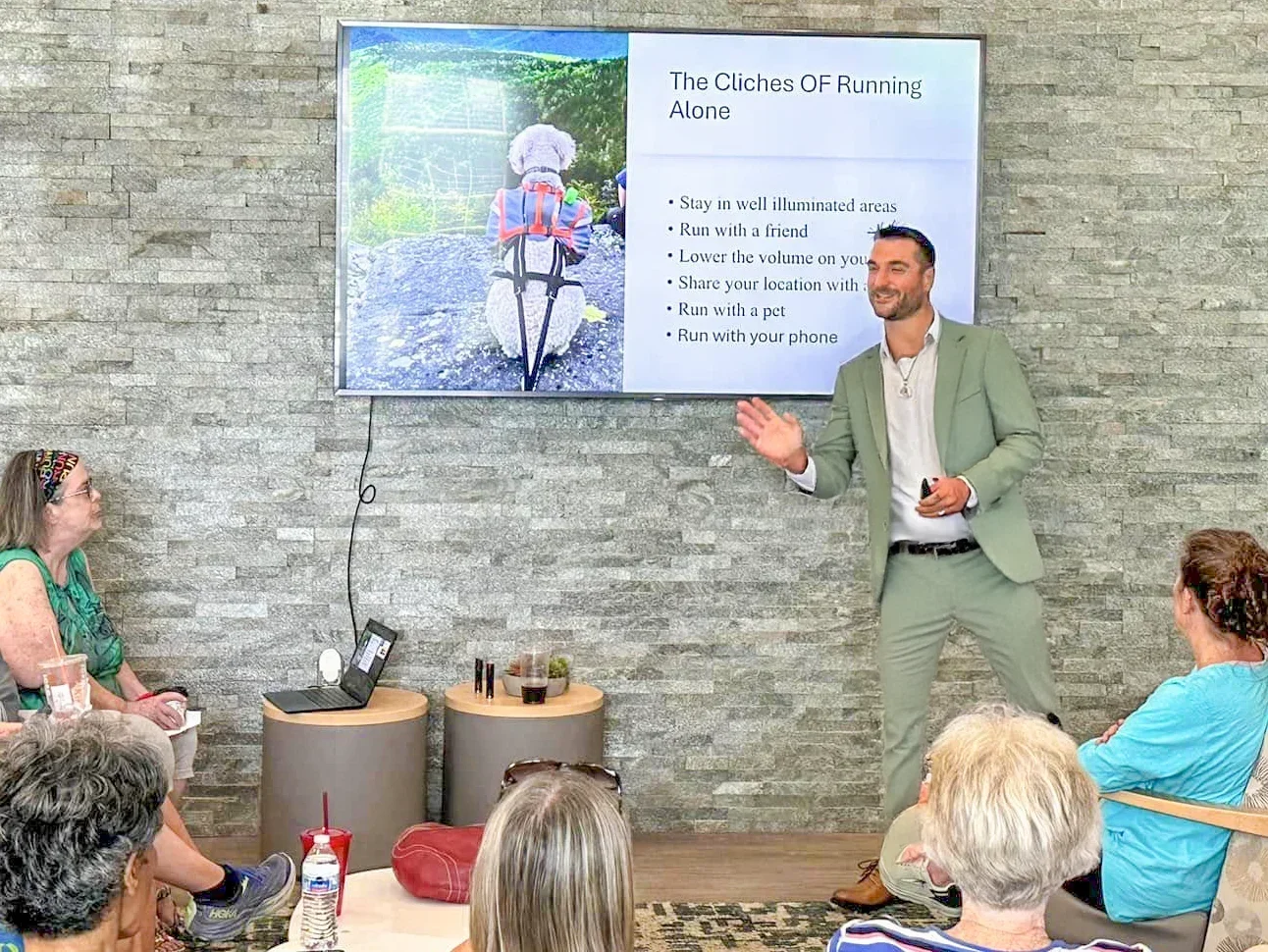 A man in a light green suit giving a presentation about running alone to a group of people seated in a room with gray brick walls. A large screen displays a hiking photo and tips for running alone.