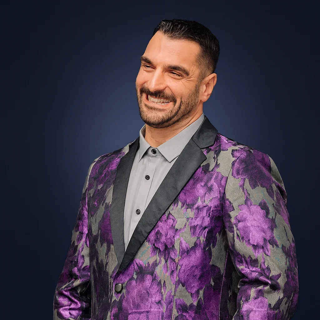 A man smiling, wearing a purple floral patterned tuxedo jacket, light gray shirt, against a dark blue background.