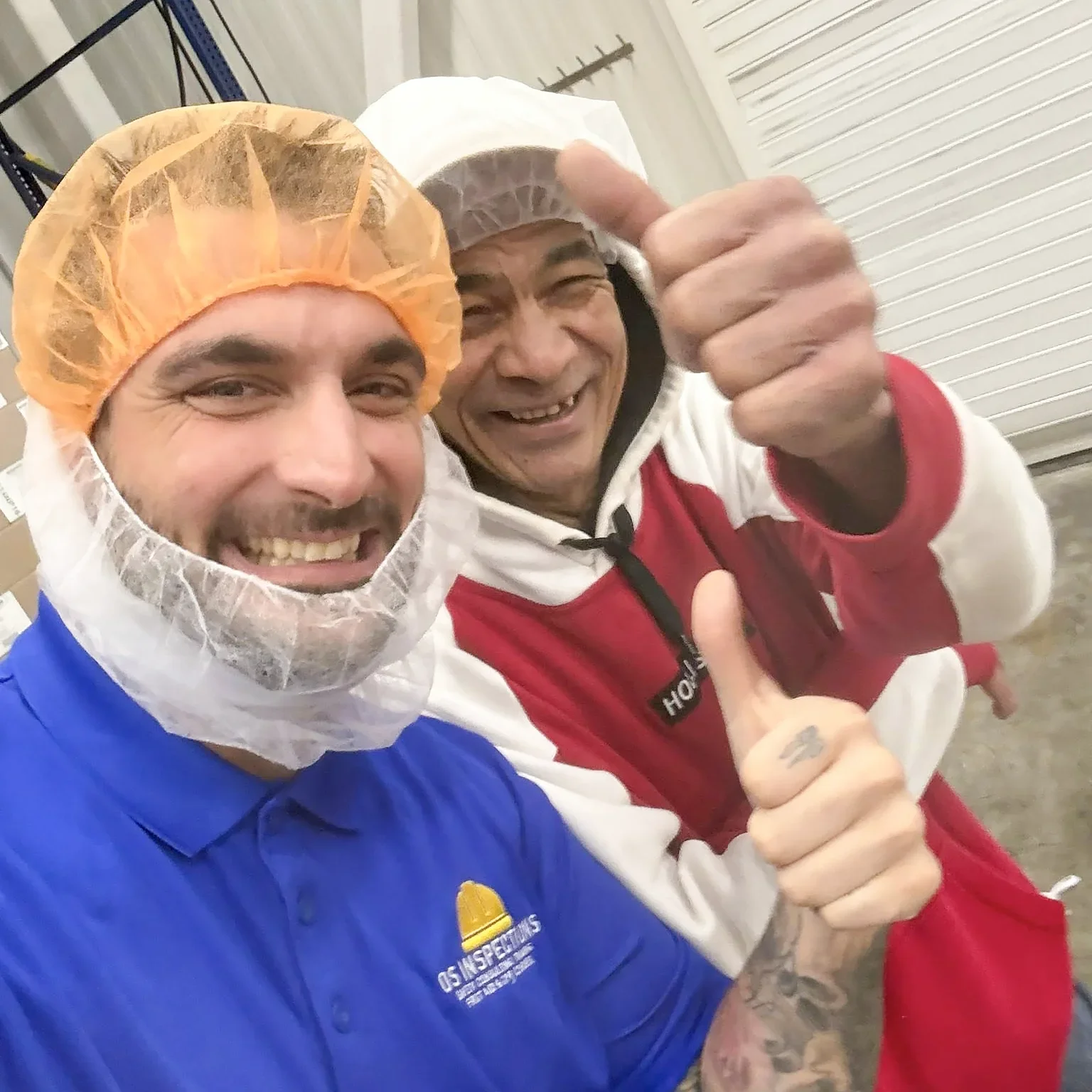 Two smiling men wearing hairnets, one in a blue shirt and the other in a red and white jacket, taking a selfie indoors, with one giving a thumbs up.