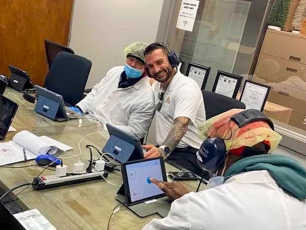 Three people sitting at a table with laptops and tablets in a room, two of them wearing headsets and one with a protective face mask, engaging with each other, smiling.