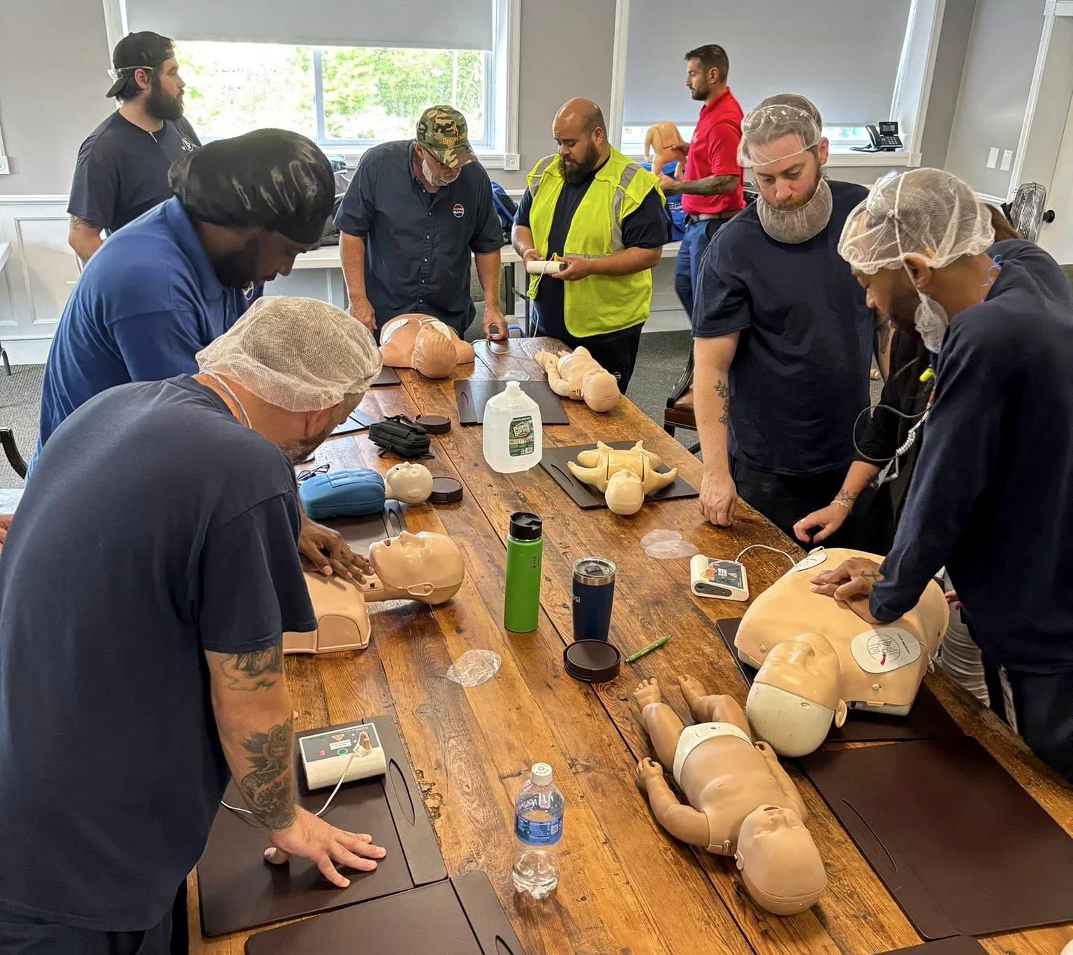 Group of people participating in a CPR training class with mannequins on a long wooden table, some practicing chest compressions and others observing, in a well-lit room with windows.