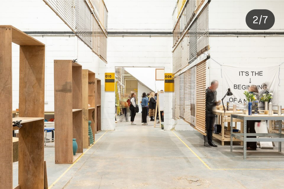People in a warehouse or workshop with high ceilings, wooden shelves, and a workstation with lamps and flowers. A banner on the wall says 'IT'S THE BEGINNING OF A'.