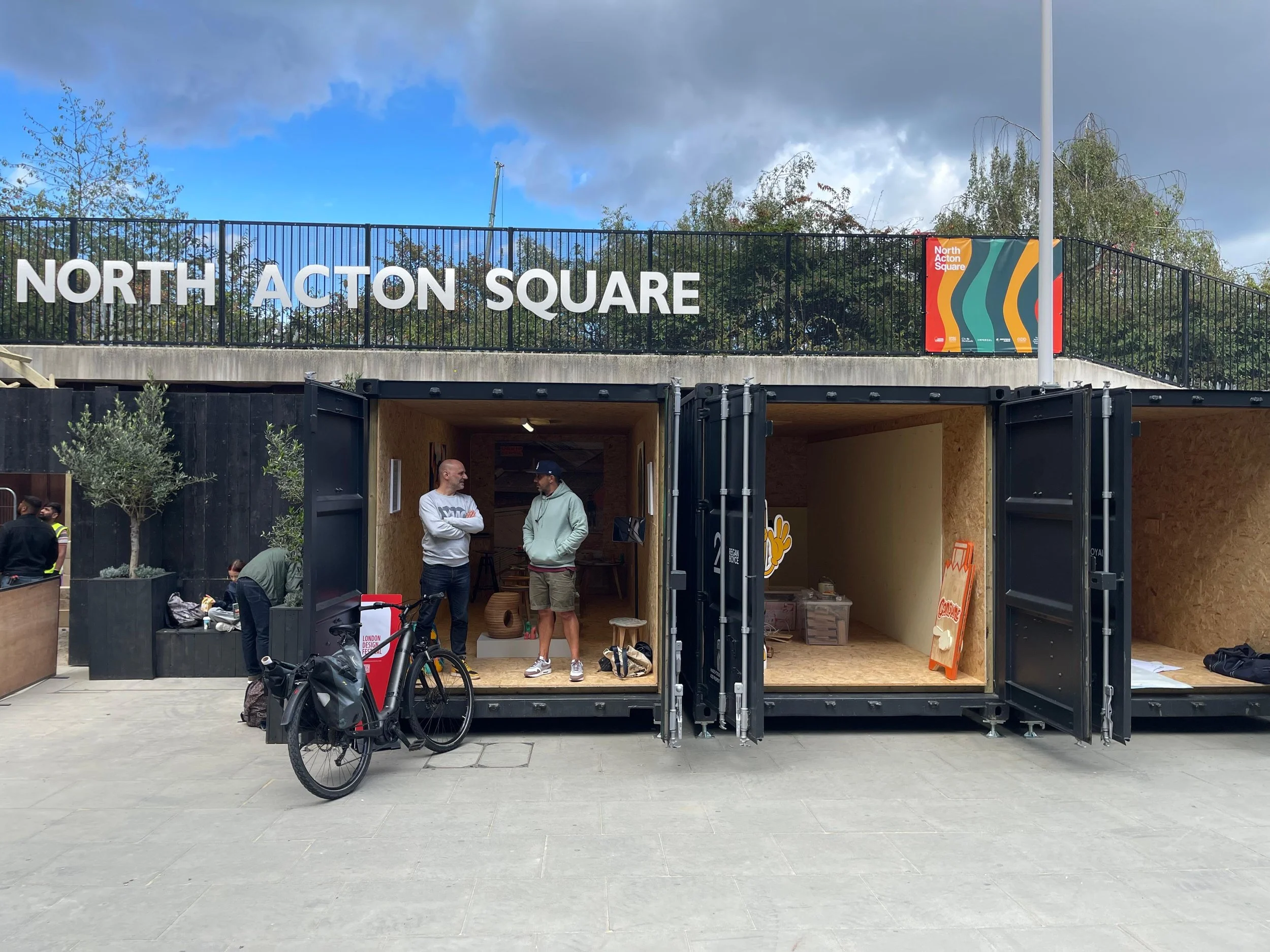 People inside and outside a mobile shop at North Acton Square with the sign visible on top, bicycles parked in front, and a cloudy sky overhead.