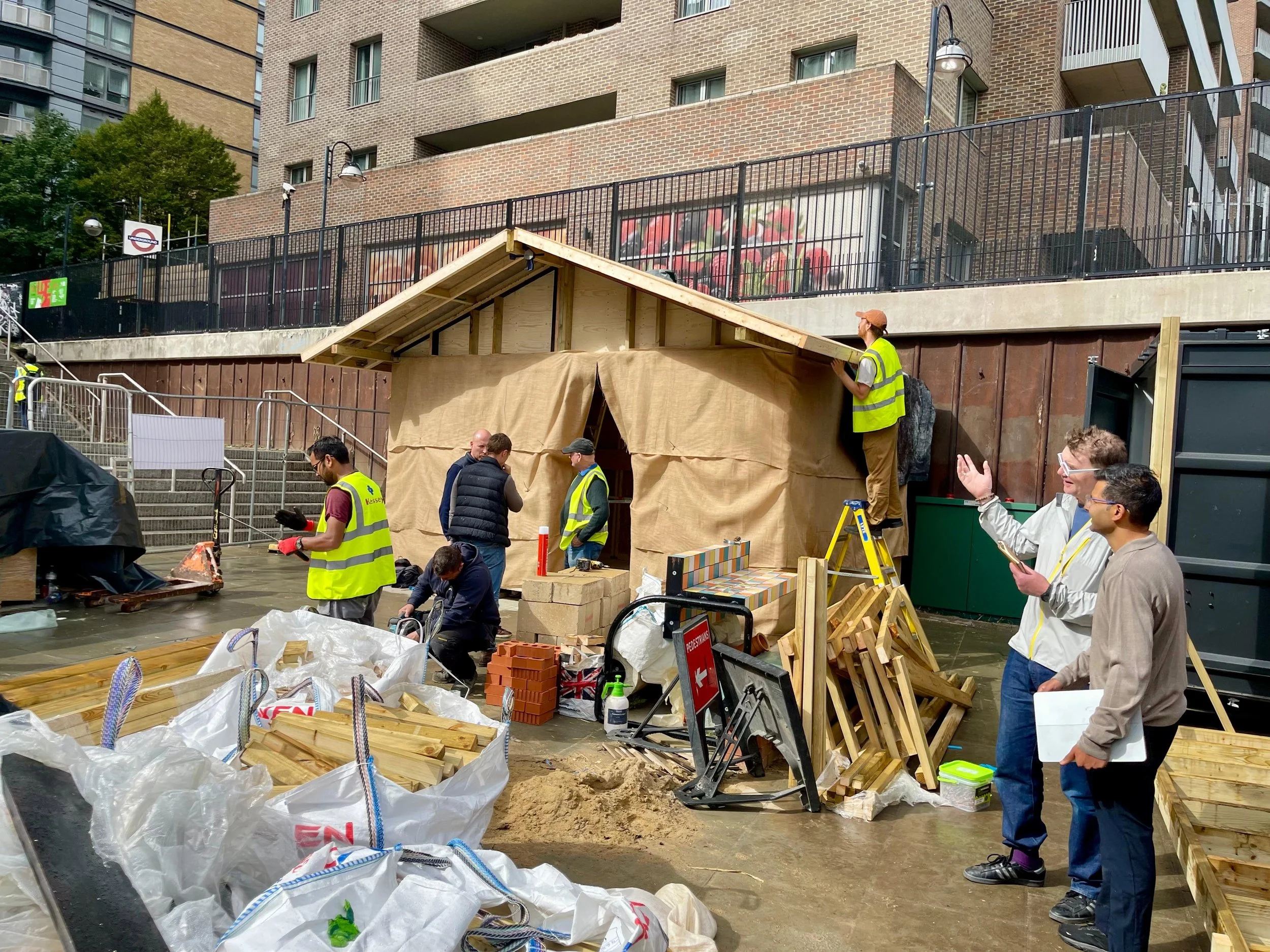 People constructing a wooden structure outdoors, some wearing safety vests, with building materials such as wood, bricks, and tools around them.