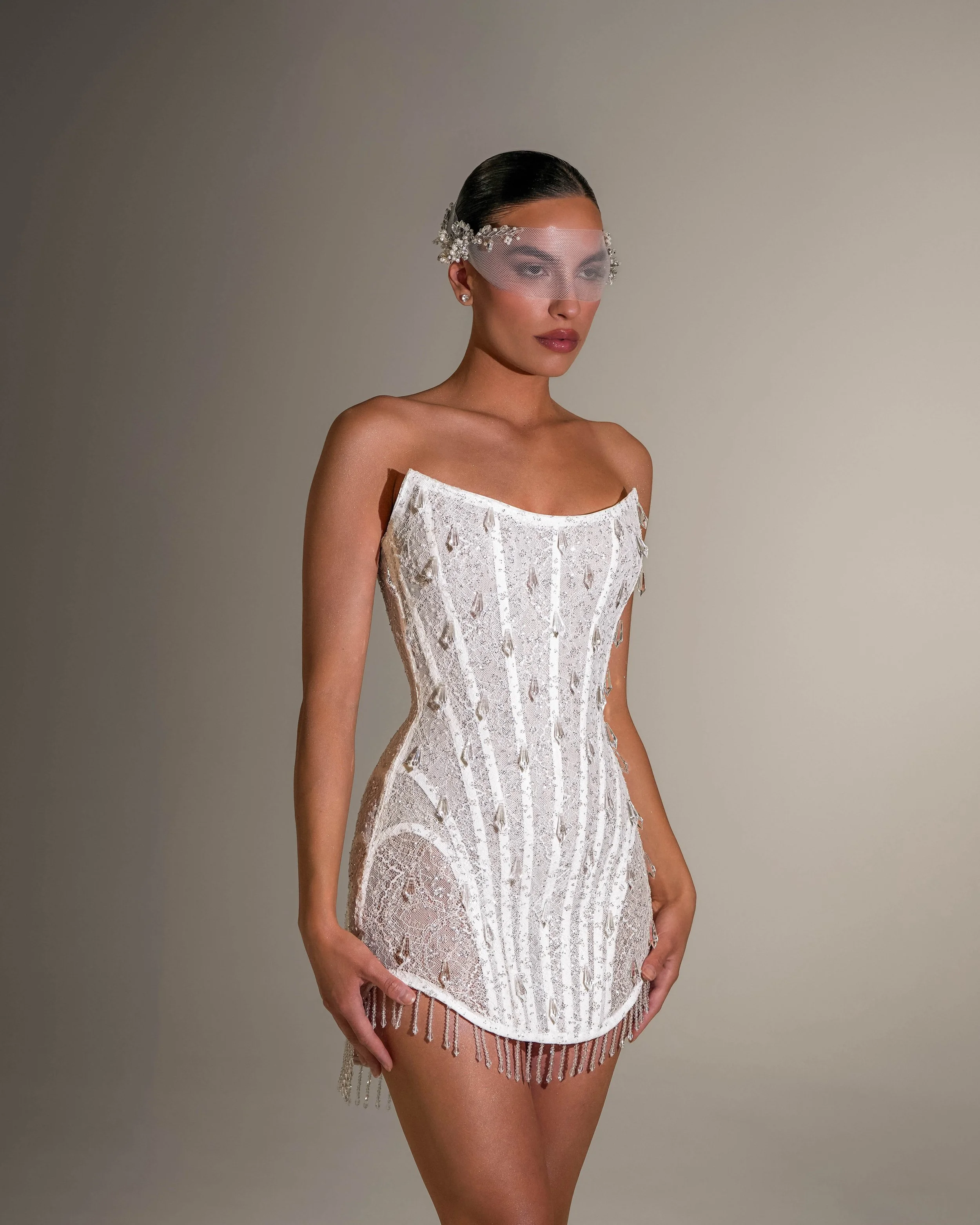 A woman with dark hair slicked back, wearing a sheer eye mask decorated with flowers and rhinestones, dressed in a white lace dress with fringe details, standing against a plain beige background.