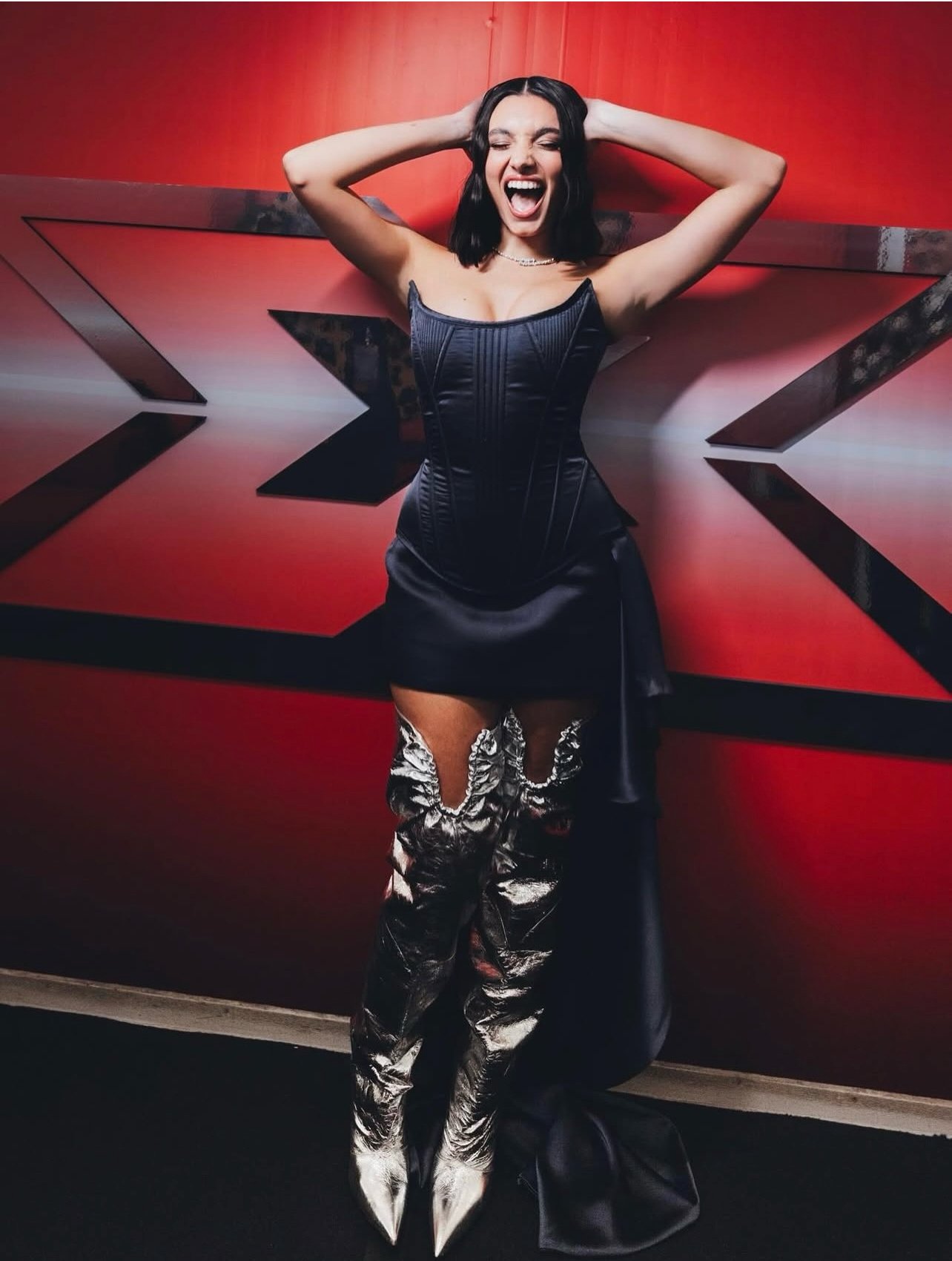 A woman wearing a black corset and shiny silver thigh-high boots, posing with her hands behind her head and smiling with her eyes closed in front of a red background with black geometric designs.