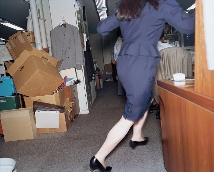 A woman in a navy suit running through an office cluttered with cardboard boxes and office supplies. OFFICE1102-O1102-RASHAAD-DENZEL.gif