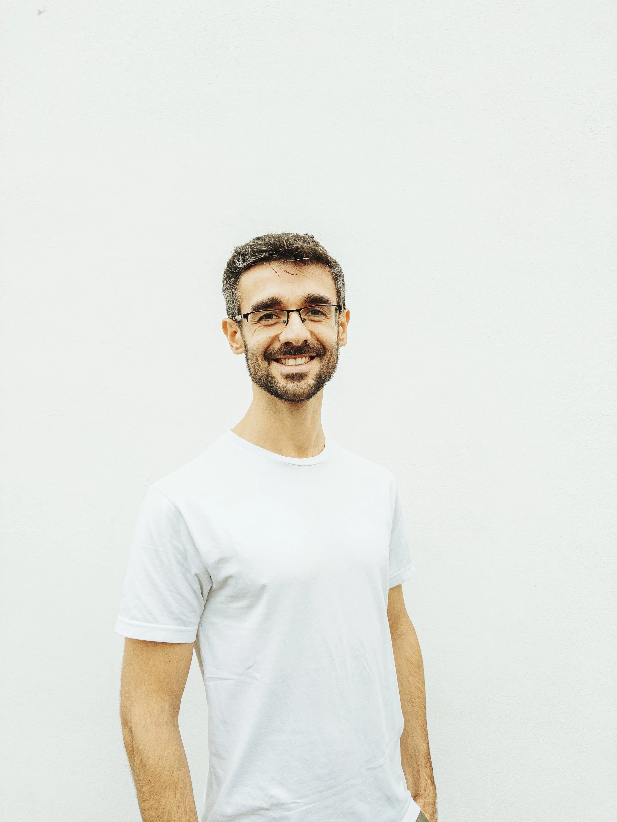 A smiling man with glasses and a beard wearing a white t-shirt, standing against a plain white wall.