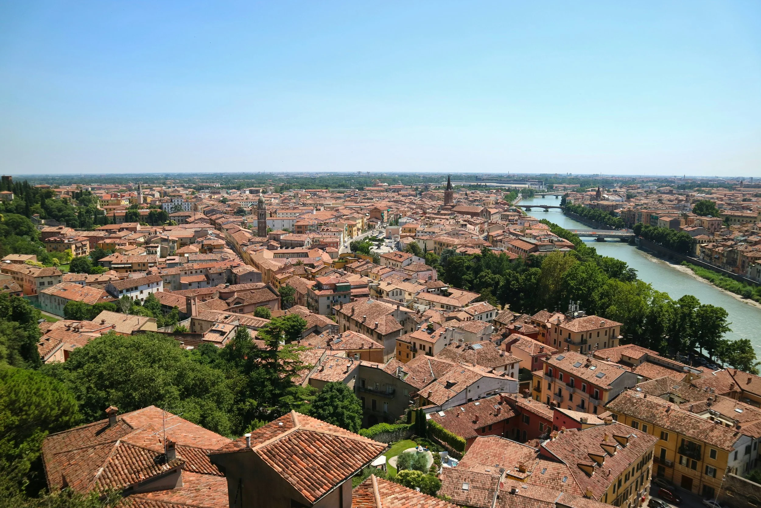 Veduta panoramica di una città con tetti in terracotta, un fiume che attraversa la città e chiese con torri. Il cielo è sereno e l'ambiente circostante è verde con molti alberi.