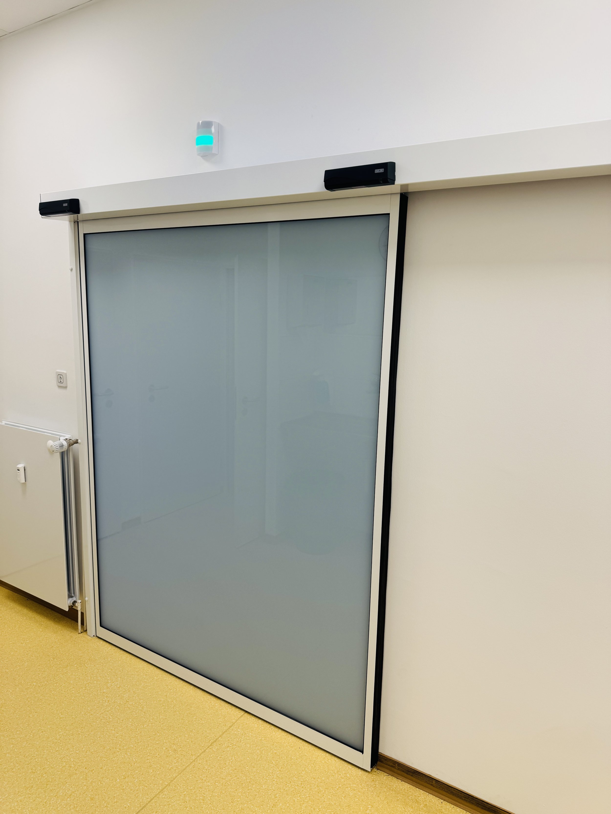 Ein verschiebbares Glas-Schiebetür im Innenraum, umgeben von weißen Wänden, mit einem schwarzen Sensor oben und einem hellgelben Boden.