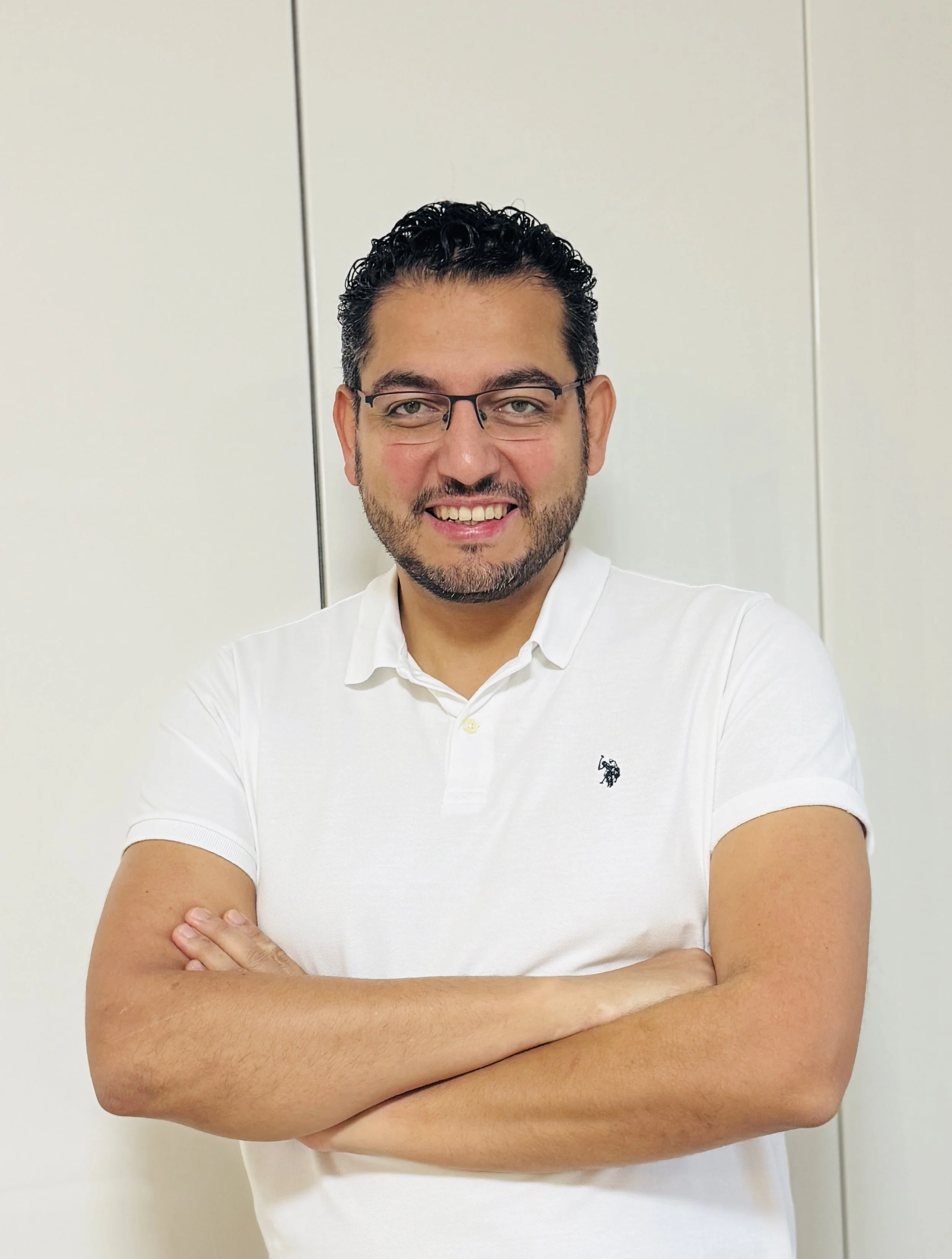 Ein lachender Mann mit schwarzen, lockigen Haaren, Brille und Bart, trägt ein weißes Polo-Shirt, steht vor einer hellen Wand mit vertikalen Schranktüren.