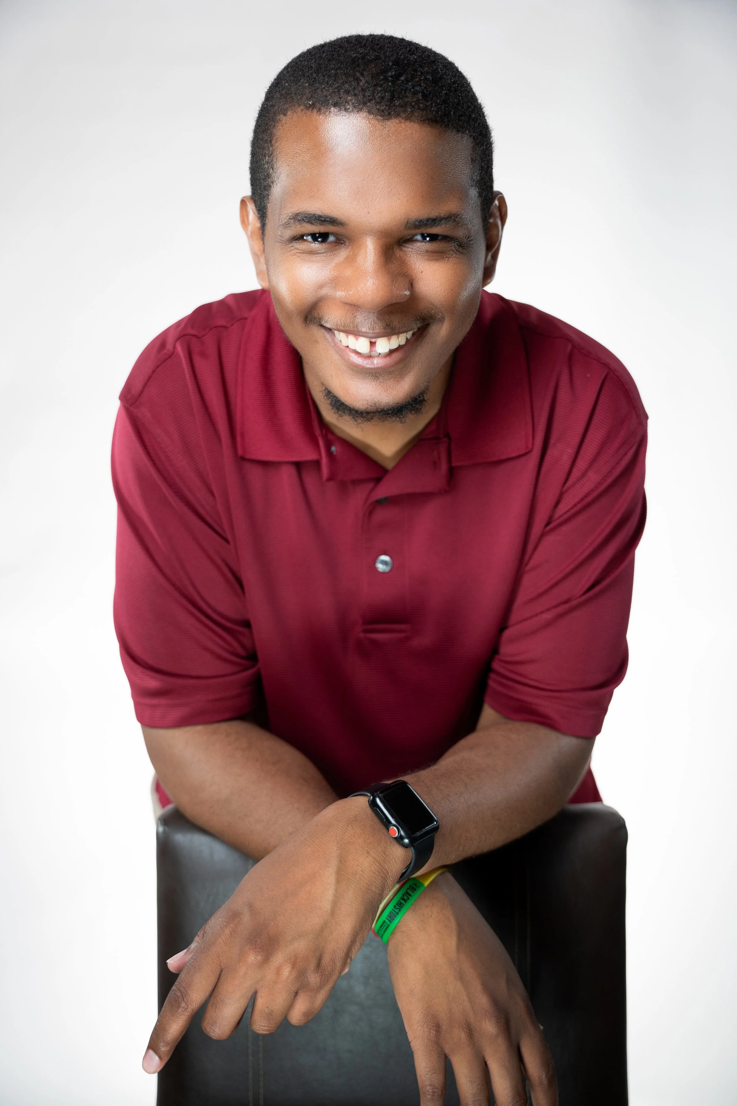 A smiling young man with short curly hair and a goatee, wearing a maroon polo shirt, a smartwatch, and a green wristband, sitting on a chair against a white background.