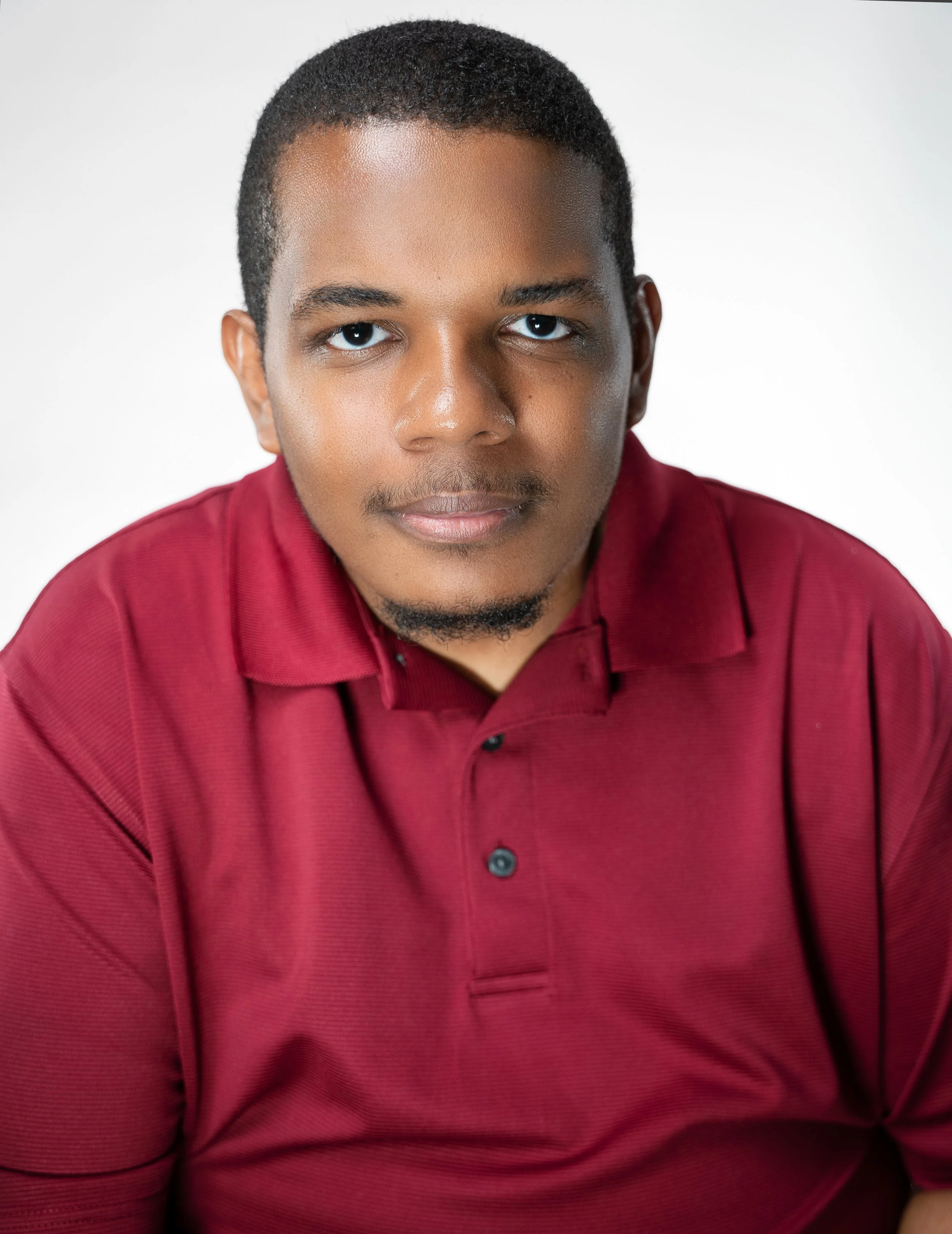 Portrait of a young man with short hair, wearing a red polo shirt, looking directly at the camera with a neutral expression.