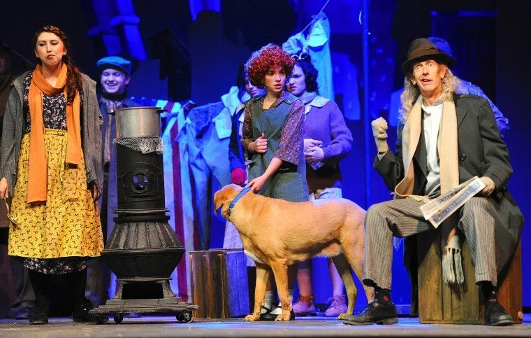 The image shows a theatrical stage scene with five actors and a dog. Two women are on either side, one with a concerned expression and the other sitting with a newspaper, wearing a hat and coat. Several actors in vintage clothing stand in the background. The scene is illuminated with blue lighting.