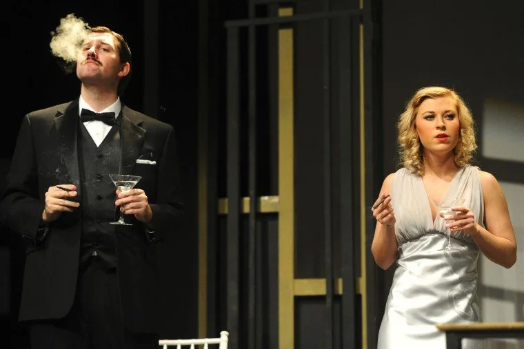 A man wearing a tuxedo with a bow tie, holding a martini glass, exhales smoke or vapor, standing beside a woman in a silver satin dress holding a drink, both appear to be at a formal event or performance.