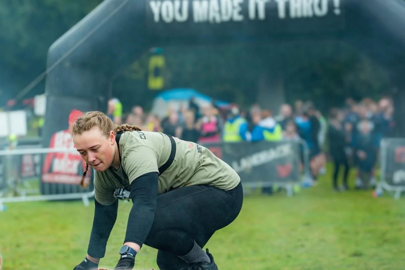 Female athlete in a green shirt and black pants is crouching on a grassy field during a race, with a large crowd and a black banner in the background.
