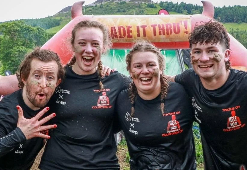 Four smiling young adults with mud on their faces and clothes, standing together outdoors in front of a large inflatable obstacle with the words "Made It Through!" on it, suggesting they participated in a muddy obstacle course or race.