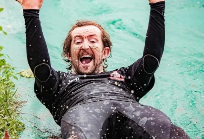 Man sliding into a body of water, smiling with arms raised, wearing a long-sleeve athletic shirt.