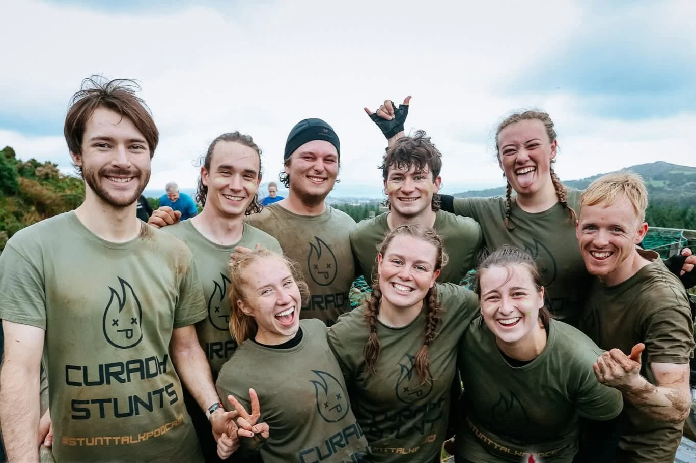 Group of smiling young people outdoors wearing matching green t-shirts with a logo and text that reads "CURADH STUNTS." They are posing happily, some making peace signs or thumbs-up gestures, with a scenic background of greenery and hills.