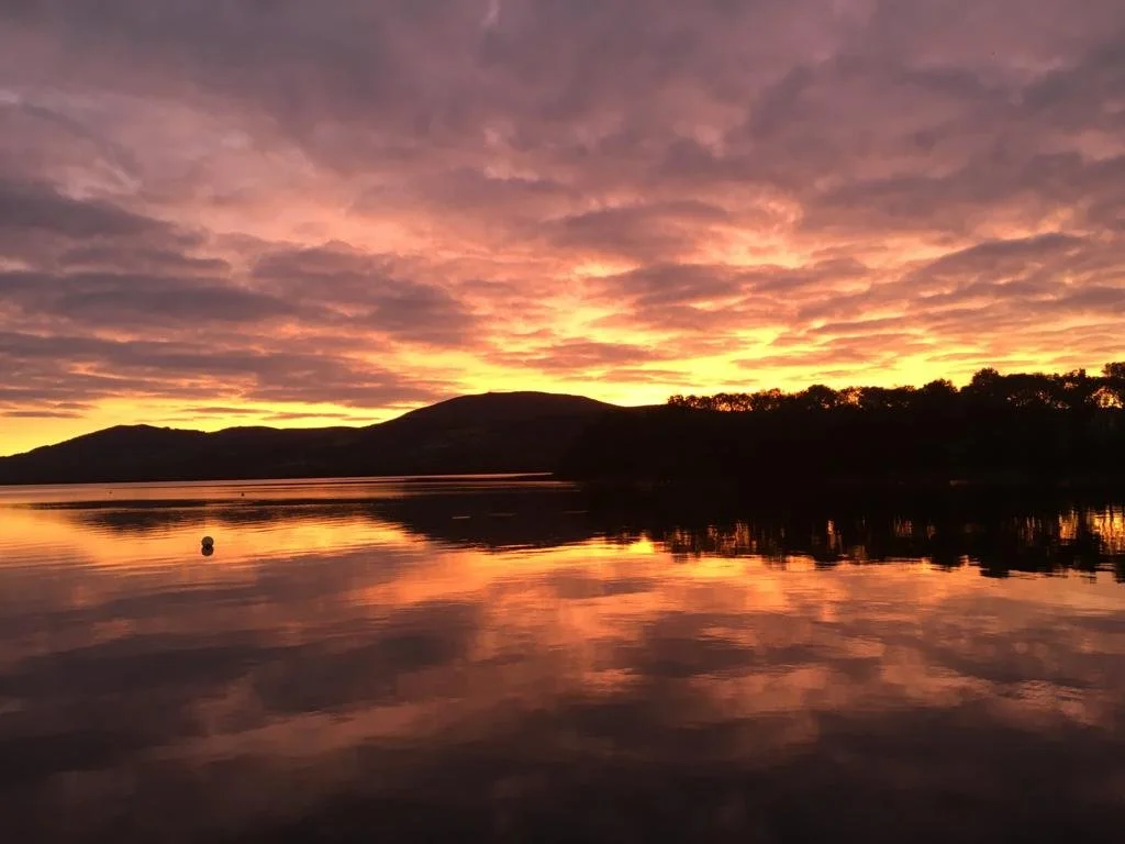 Sunset over a lake with reflections, a distant silhouette of hills, and a partly cloudy sky with pink, purple, and orange hues.