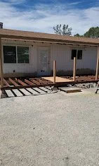 Front view of a house under construction with wooden deck framework in progress, gravel driveway, and a clear sky.