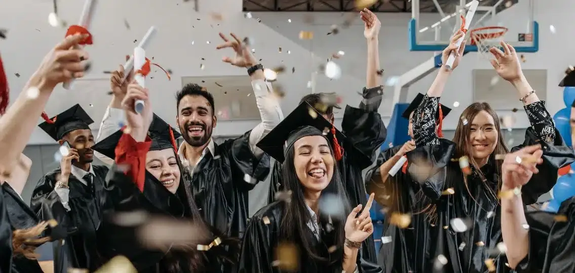 Group of graduates in caps and gowns celebrating at a graduation ceremony with confetti and joyful expressions.