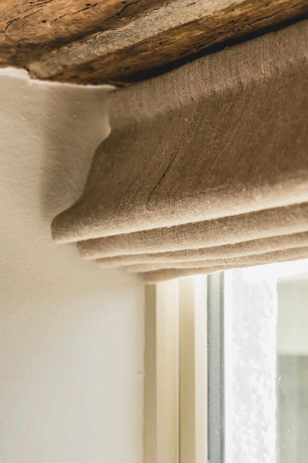 Close-up of rustic-looking beige fabric roman blind hanging in window, with a glimpse of a cream wall and window frame in the background.