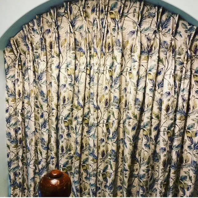 View of decorative oval window showing handmade pleated curtains with a floral and leaf pattern in shades of blue, green, and beige.