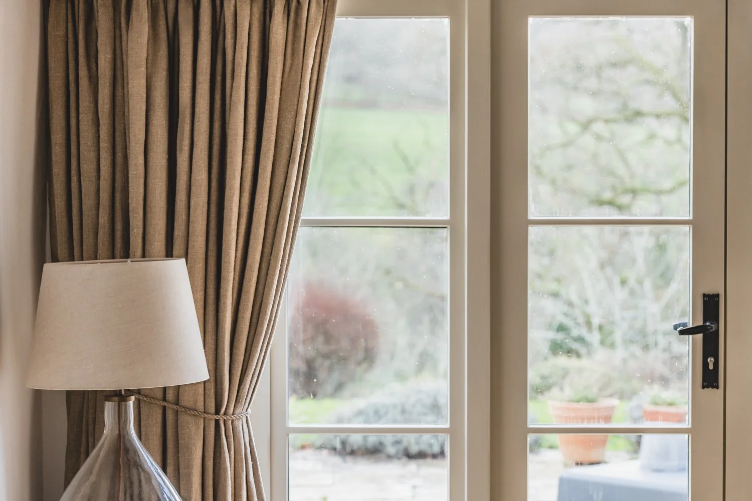 A glass door with beige curtains tied back on the left, outside there are potted plants and greenery visible through the glass panels.