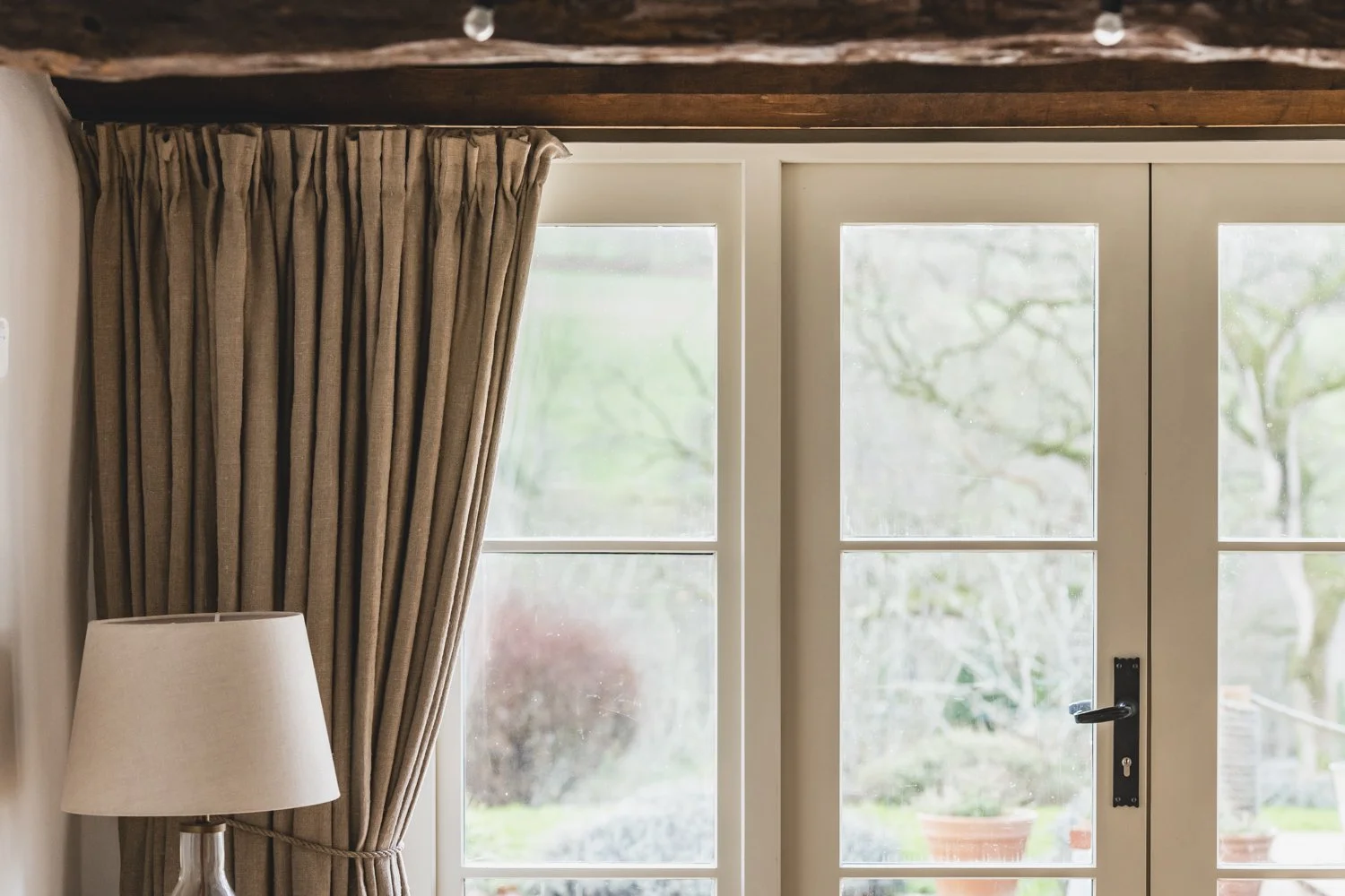 Interior view of a room showing a closed white wooden door with glass panes, luxury beige curtains on the left side, and a white table lamp with a beige lampshade.