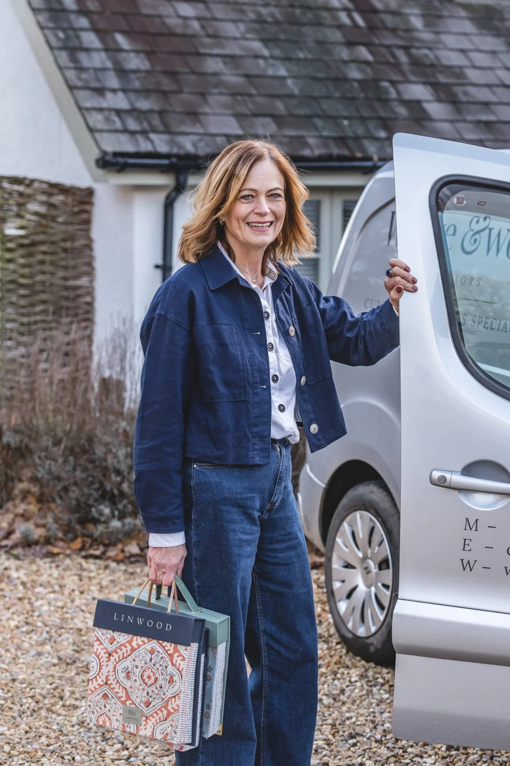 A woman stands outside a cottage bungalow, smiling, holding a fabric book with one hand and the door of a silver van open with the other. She is dressed in a white shirt and a dark blue jacket, blue jeans, and stands on a gravel driveway.