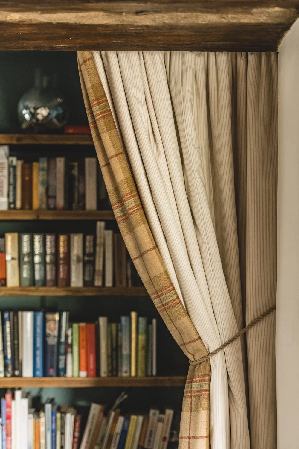 A partially drawn weighted cream-coloured fully lined curtain, covering a bookshelf filled with various books.