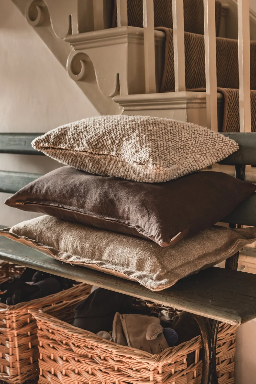 Stacked decorative pillows on a distressed wooden bench in a cosy interior, with a staircase in the background and wicker baskets beneath the bench.