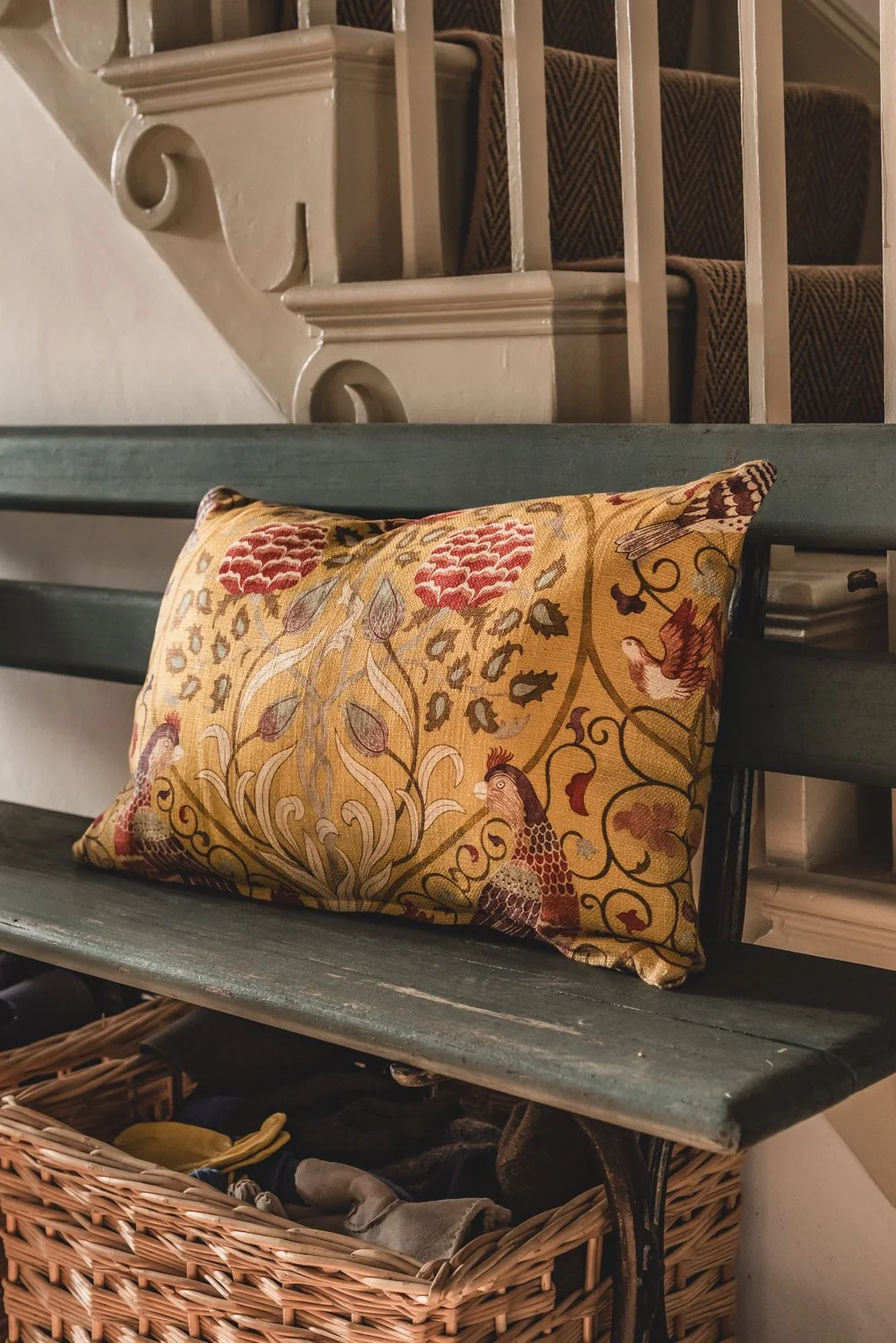 A decorative pillow with intricate floral and bird patterns resting on a dark green wooden bench, with a staircase with brown patterned carpet and a white banister in the background, and a wicker basket filled with folded fabric underneath the bench.