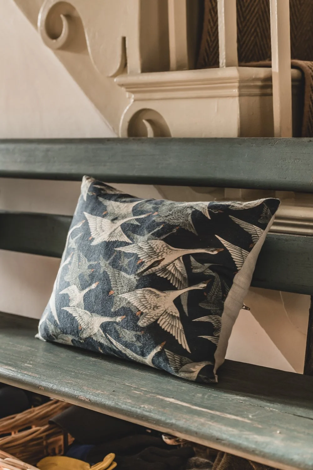A decorative pillow with bird illustrations resting on a green wooden bench underneath a staircase.
